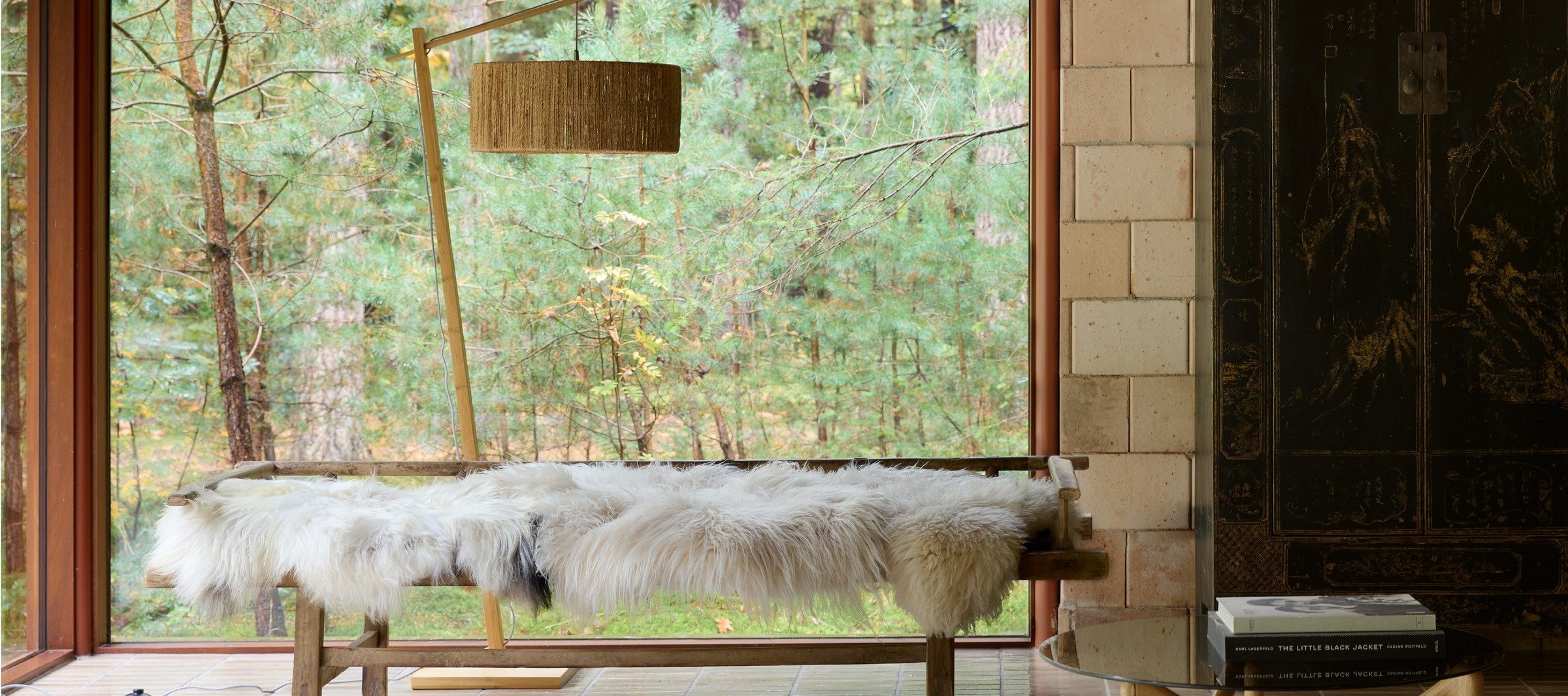 A sheepskin-draped vintage bench beneath a rattan pendant lamp, forest view behind